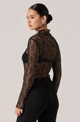 A woman is wearing a sheer black long-sleeve top with a high ruffled collar. The top is decorated with small, shiny, white polka dots. She pairs it with high-waisted black pants.